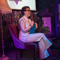 A white woman with short brown hair and black frame glasses, wearing a sleeveless white pin stripe suit and sat on a purple chair on stage