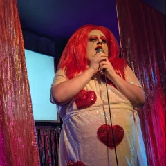 A drag artist wearing a red wig and a white dress sequin dress with red red sequin hearts on it singing into a microphone