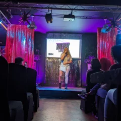 A white woman with blonde hair performing on stage singing into a microphone wearing a black and white striped top and light denim skirt and black boots.