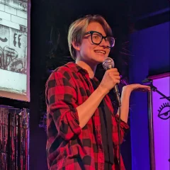 A woman with short light brown hair and large frame glasses wearing a red and black checkered shirt speaking into a microphone