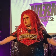 a Drag Artist with red hair and sparkly black sequin dress with a heart embellishment that reads "glitter king".